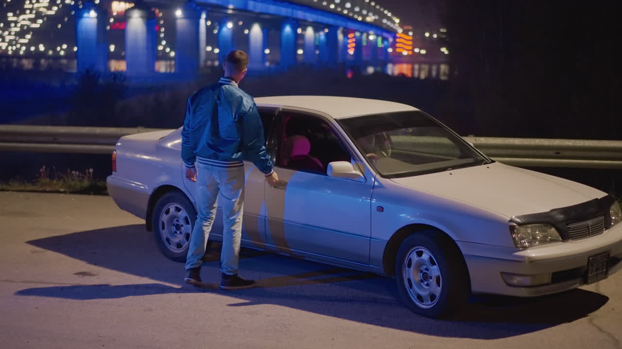 un hombre blanco joven se acerca a un coche aparcado bajo las luces de neón de un puente; tenso intercambio nocturno cuando el hombre abre la puerta y se sienta en el sedán, luces azuladas y rosadas, ambiente urbano al borde de la carretera, reflejos