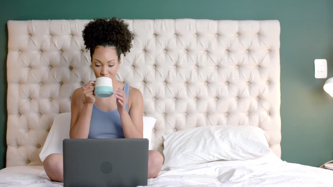 Donna birazziale seduta sul letto che beve caffè e usa il laptop in camera da letto, al rallentatore.