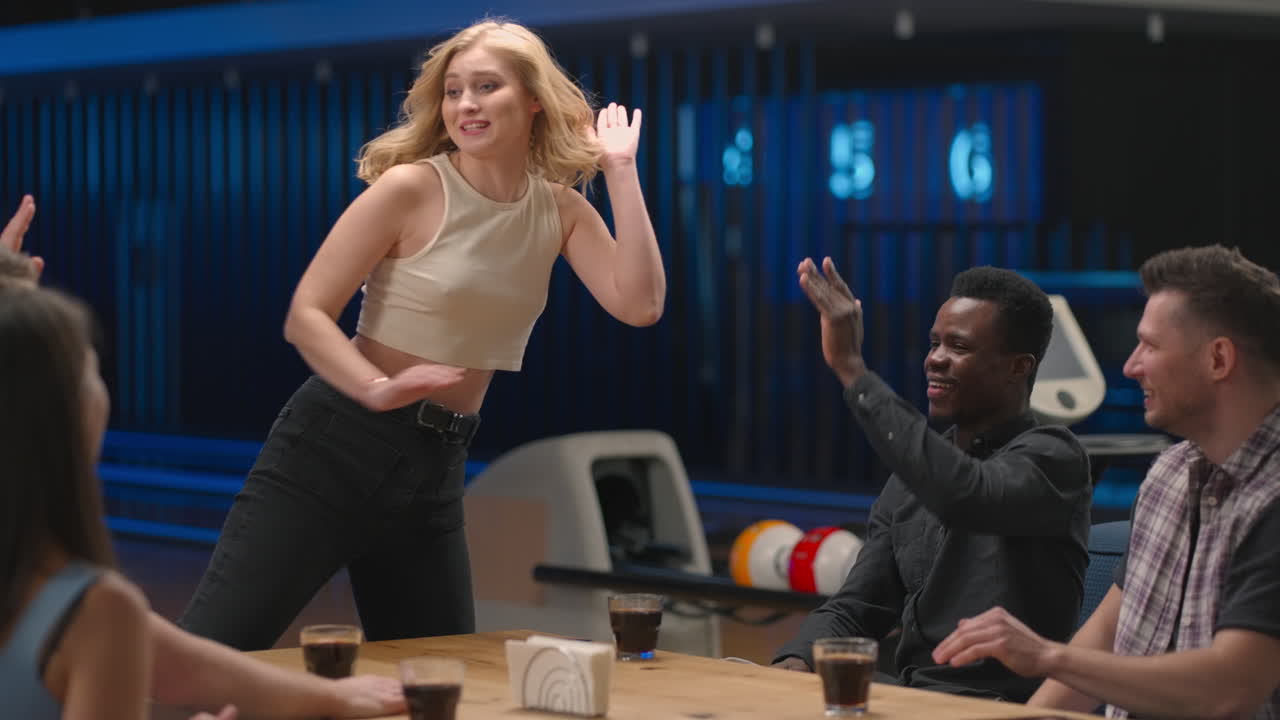 A joyful blonde woman runs up to friends giving five men and women sitting at a table in a bowling club. Celebrate victory and streak. To cut and congratulate on the victory. Team success.