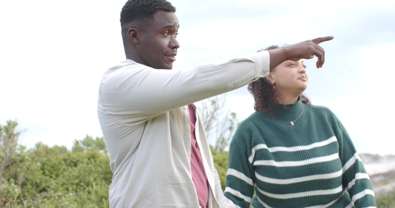 Couple afro-américain se tenant la main, marchant sur un sentier herbeux, le partenaire de gauche pointant vers les dunes.
