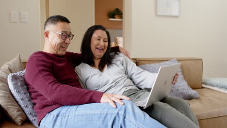 senior asian couple relaxing on sofa, enjoying laptop together at home