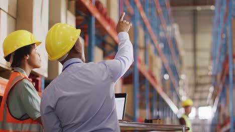 Trabajadores-Afroamericanos,-Hombres-Y-Mujeres,-Con-Casco-Y-Usando-Una-Computadora-Portátil-En-El-Almacén