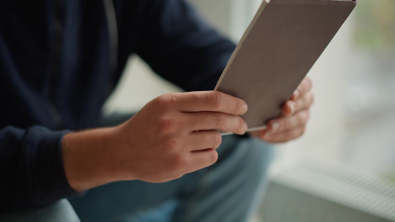 人物が両手で開いたノートを手に持ち、調べているクローズアップ。手の位置と本の向きの相互作用に焦点を当てています。シーンは窓の近くの屋内で、柔らかな自然光が差し込んでいます。