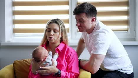 blonde woman sits in armchair swinging her newborn baby. husband sits beside admiring his son.