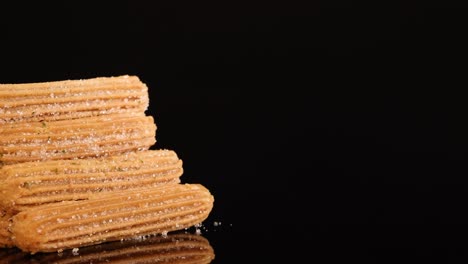 a neatly stacked pile of churros rotates smoothly on a glossy black surface, illuminated by soft studio lighting, highlighting texture and sugar crystals