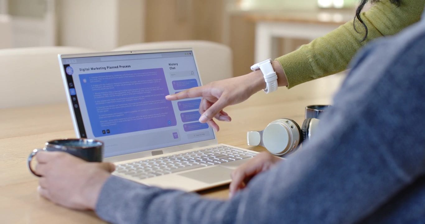 Partner reaching in and tapping laptop at table, marking plan, seated moving hand while holding mug