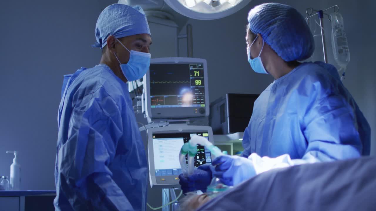Mixed race surgeons wearing protective clothing looking at heart monitor in operating theatre