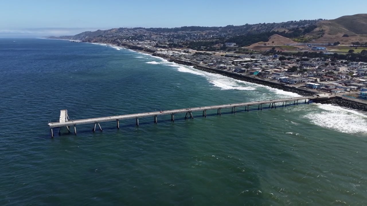 Imágenes de dron matutinas del Muelle de Pacifica y la costa de California. Océano azul cristalino, olas suaves, colinas iluminadas por el sol y un pueblo costero con una composición aérea dramática. Ideal para proyectos paisajísticos