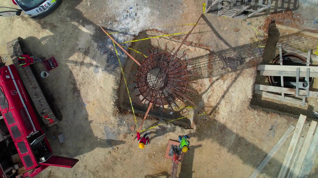 High-angle aerial drone top-down shot rising rapidly and pulling back over a steel rebar reinforcement cage inside a foundation shaft at a civil engineering construction site