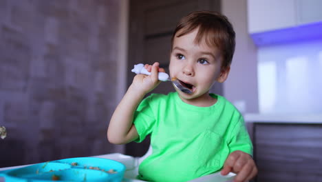 adorable little toddler sits holding a spoon in his mouth. mom holds a full spoon prepared for her son. blurred backdrop.