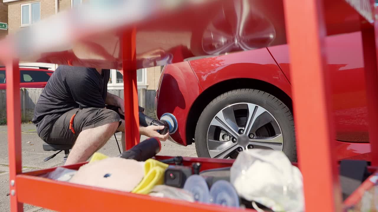 Polishing rear bumper of red car whilst looking through tool trolly