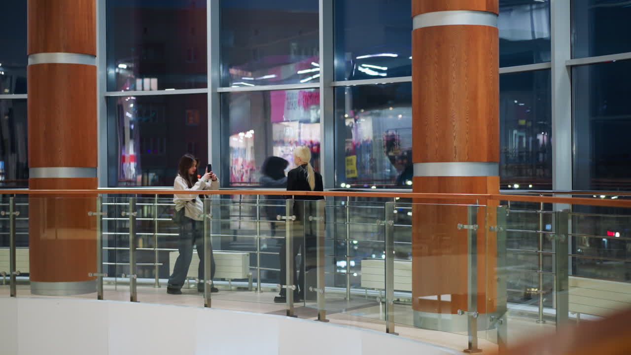 Girl holding phone capturing photo of her friend indoors near large glass window while colorful city lights reflect on glass creating bright vibrant atmosphere in modern shopping mall during evening