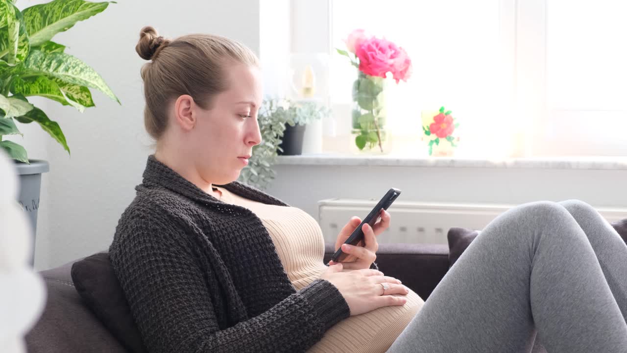 Pregnant white woman sitting on the couch typing on the phone with the hand on the belly slowmotion at daylight