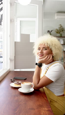 vertical video: smiling woman enjoying coffee and donut at cafe table, smartwatch on wrist