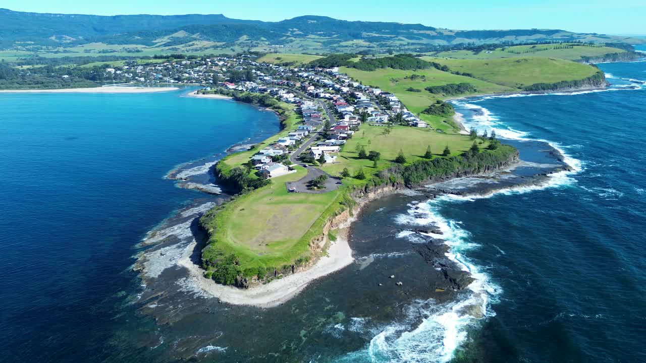 Drone aerial landscape of ocean waves crashing against rocky coastline reef with suburban town housing and residential neighbourhood homes on headland in Gerroa South Coast Australia travel holidays
