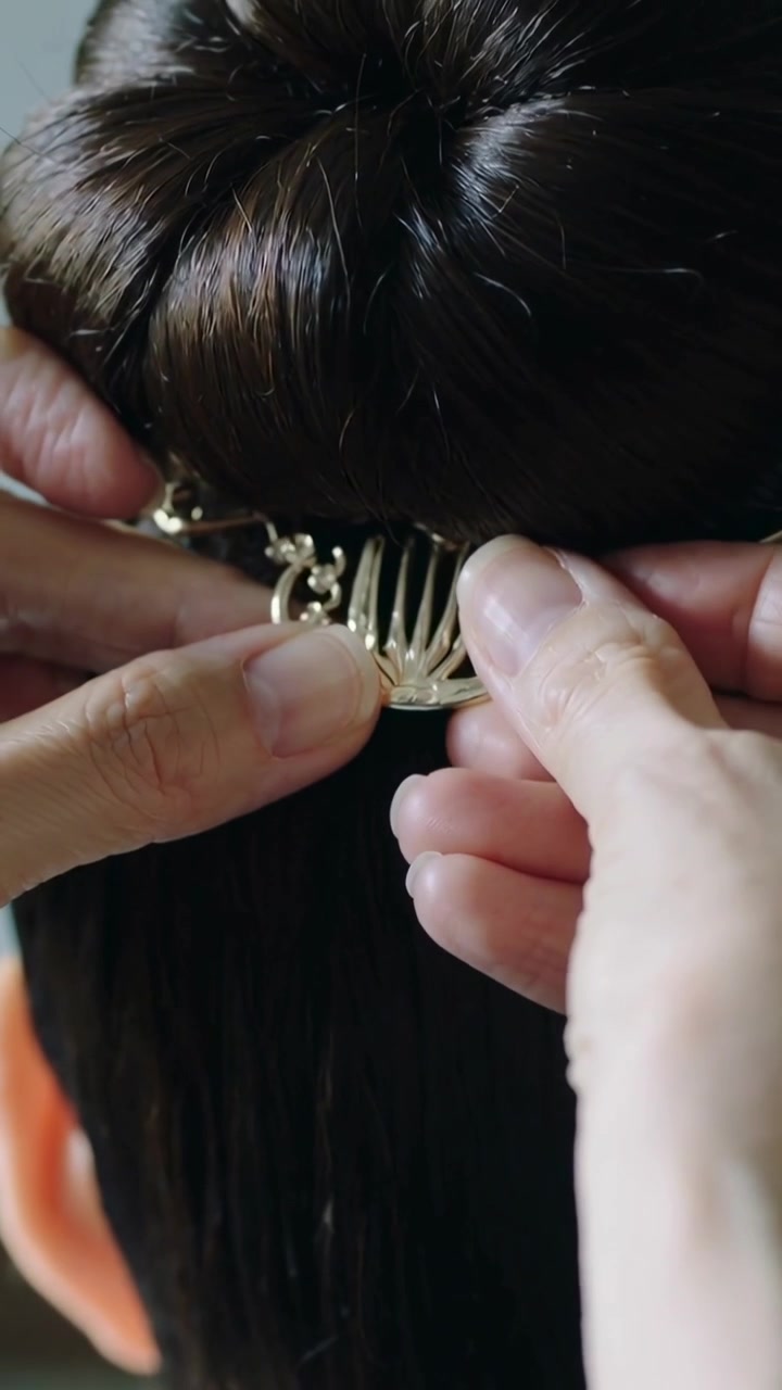 Video verticale: Mani di una stilista che portano un pettine d'oro, lo fanno scivolare in uno chignon in un salone per fissare una catena