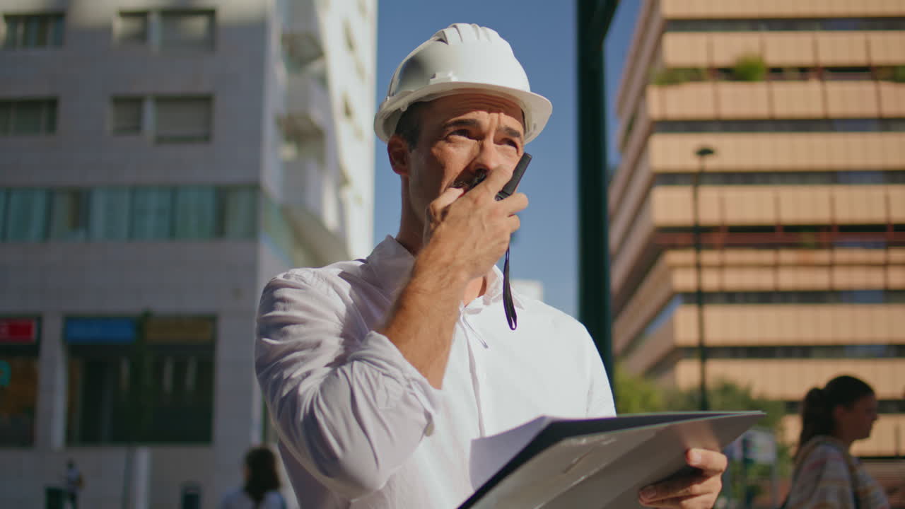 Serious builder watching plan at sunbeams city closeup. Man using walkie-talkie