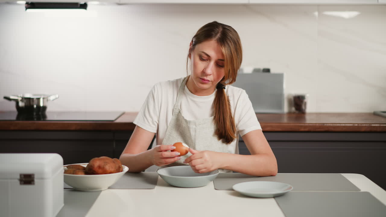 Housewife wearing white shirt and apron sitting at kitchen table focused on peeling shell off cooked egg over bowl, with potatoes and kitchen items arranged neatly on clean countertop in modern home
