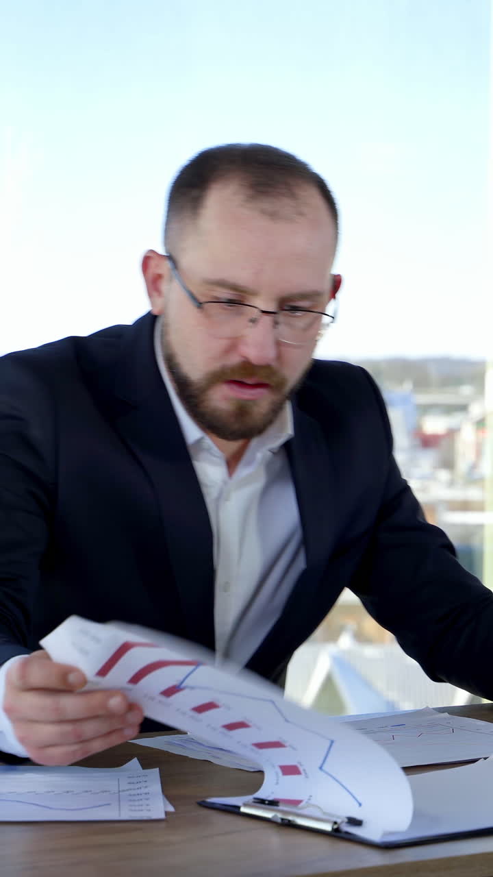 Nervous businessman at office. Office worker sitting at his desk dispirited of some difficulties. Angry man in suit scattering papers at his workplace. Vertical video