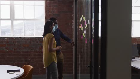 Diverse-male-and-female-business-colleagues-wearing-face-masks-brainstorming-pointing-to-glass-wall