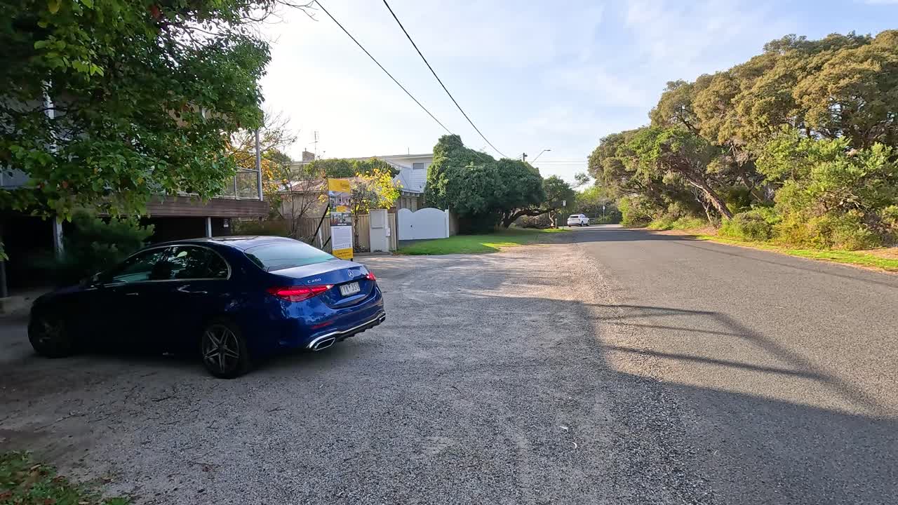 A blue car exits a driveway and drives down a sunlit residential street in Point Lonsdale, Victoria