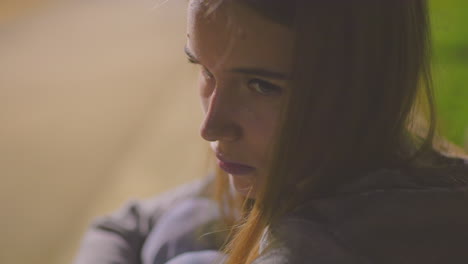 close-up of tired young woman lifting her head slowly, showing signs of exhaustion and deep thought, surrounded by soft blurred background with warm tones of an evening park setting