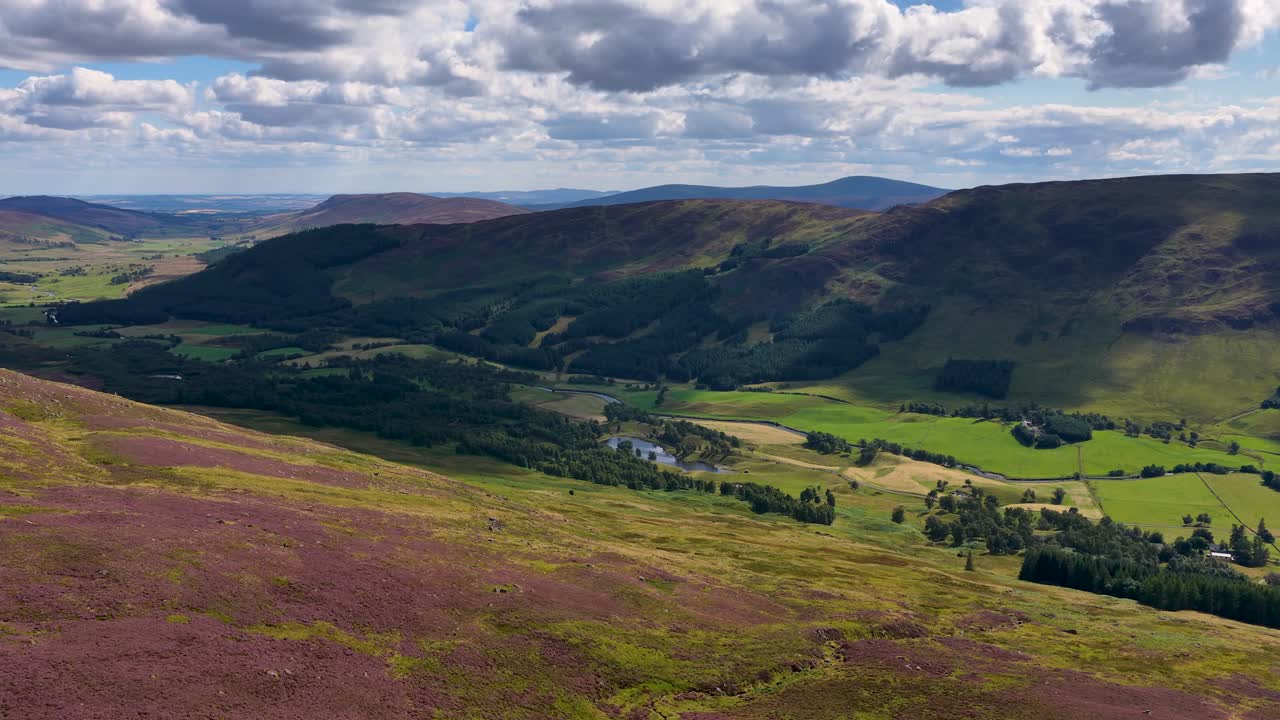 Drone camera slowly pans across vibrant heather hills, green valleys, and dramatic cloud shadows