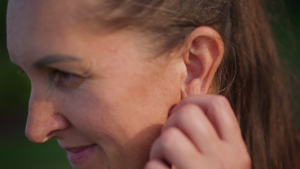 Signora pensierosa che si tocca l'orecchio all'aperto al tramonto. Donna pensierosa con la coda di cavallo che sorride dolcemente al tramonto. Primo piano di una donna con la coda di cavallo che si sistema l'orecchio in un'atmosfera nostalgica all'aperto al crepuscolo