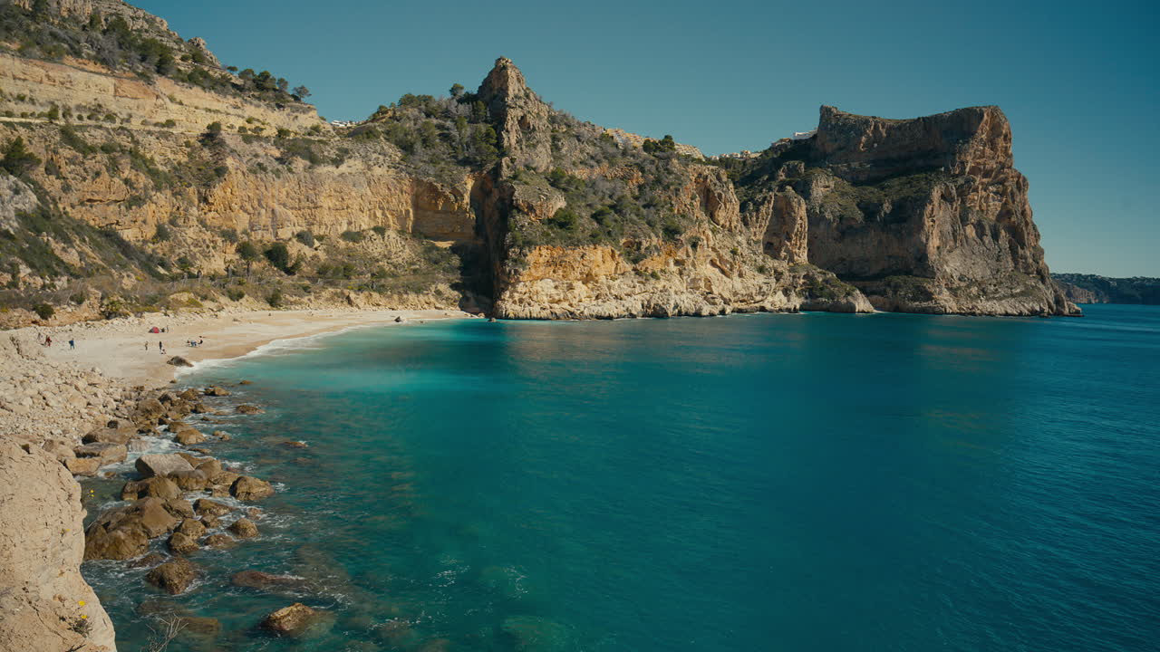 Stor middelhavsbugt med turkisblåt vand, dramatiske klipper og små strandgæster langs kysten, der tilføjer skala, sommerstemning og en indbydende kystflugt