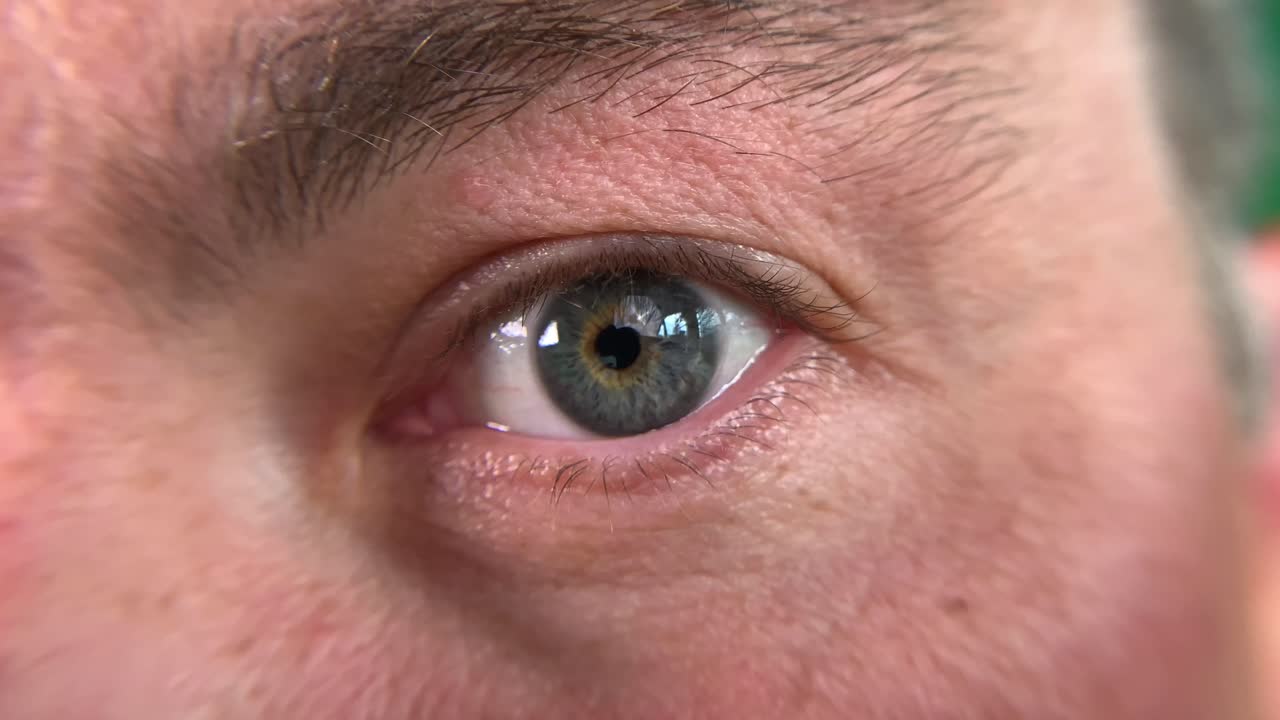 Interior, close up of mans left eye, staring straight ahead. He has brown thick eye brows and blue eyes with hazel