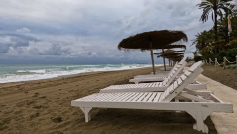 toma 4k de una playa vacía en un día nublado con hamacas y sombrillas de paja en marbella, españa