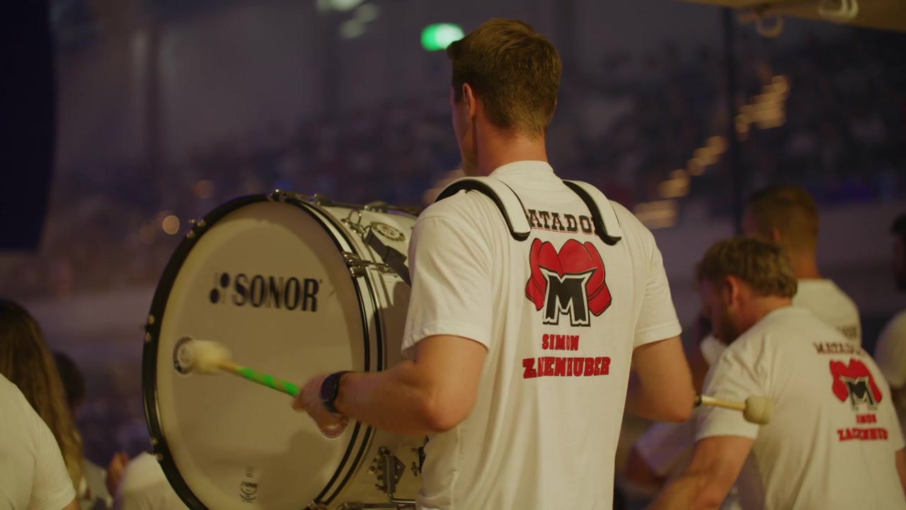 Trommeslagere sætter gang i publikum ved et live indendørs sportsarrangement. En passioneret fangruppe spiller højt til støtte for deres hold. Filmet inde i en fyldt arena