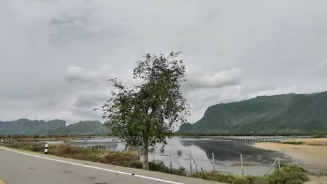 Conduciendo-Por-Una-Carretera-Junto-A-Un-Lago-Con-Colinas-Montañosas-Al-Fondo-En-Tailandia