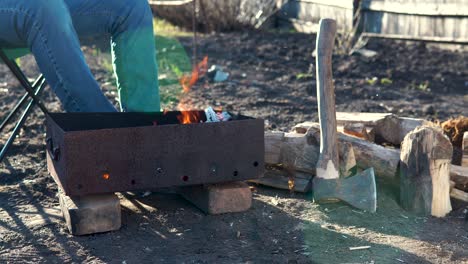 an unrecognizable man relaxedly sits on a chair in front of a bonfire in the garden. a young man is resting in the backyard, preparing a barbecue.