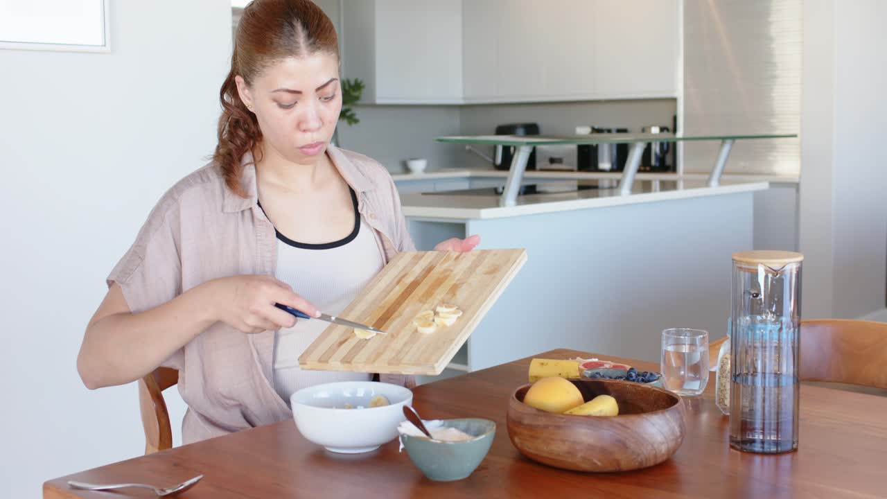 African American woman at table slicing banana, angling board, pouring rounds into bowl for snack