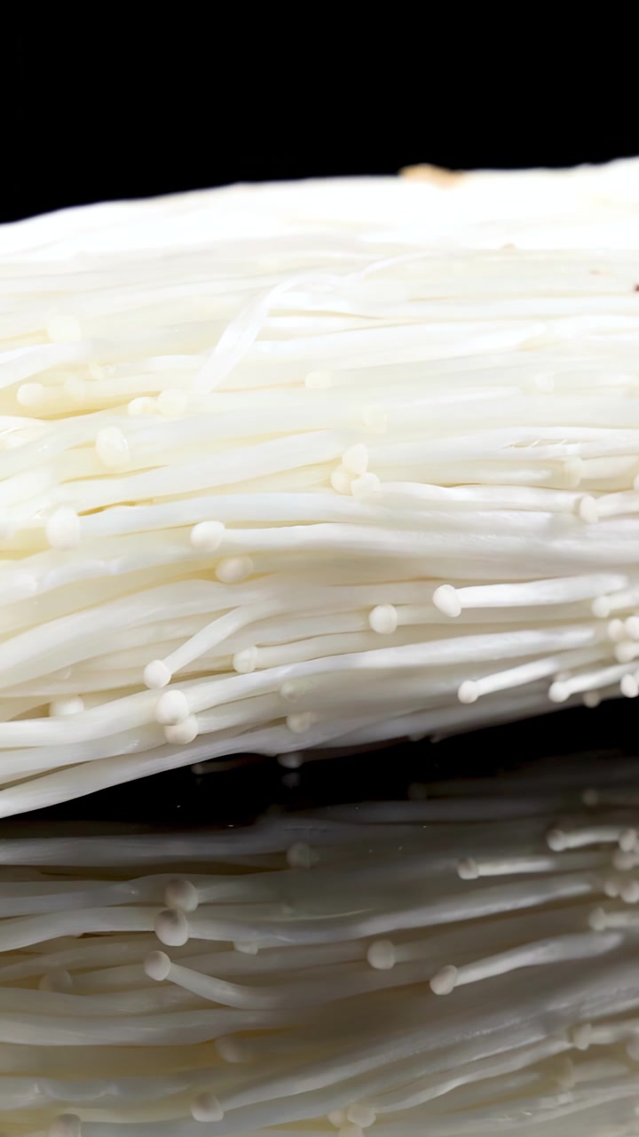 A bundle of enoki mushrooms slowly rotates on a glossy black reflective surface under bright studio lighting, revealing fine details and smooth textures