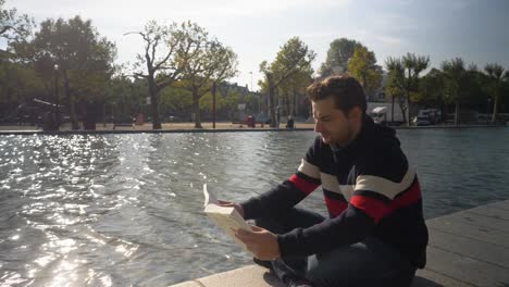 Nice looking young man sitting by a canal in Amsterdam concentrated reading a book