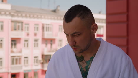 Upset-man-with-tattoos-on-body-in-bathrobe-stands-on-terrace