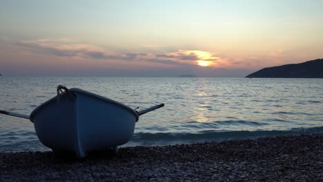 a lone row boat sits on a pebbly beach as the sun sets behind it and gentle waves roll in