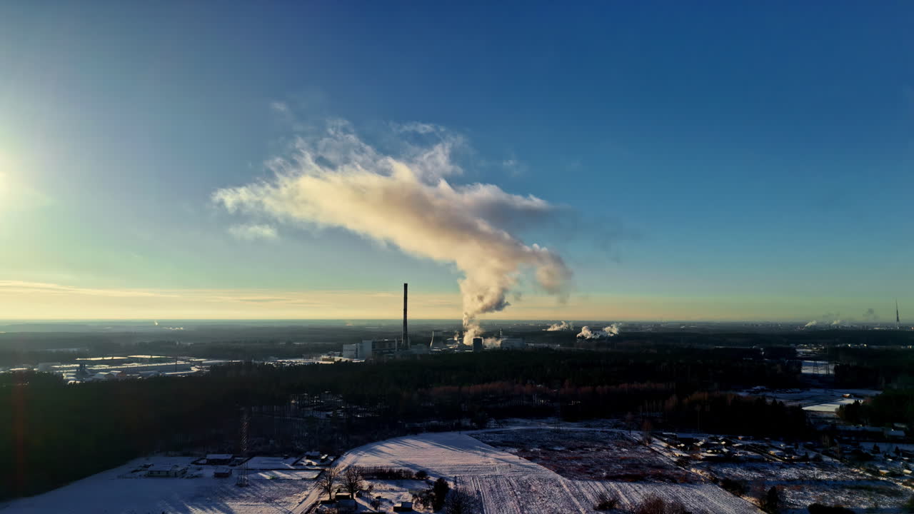 雪の降る田園地帯をドローンで空撮。発電所の煙突からは長い蒸気の煙が澄んだ青空に放出され、冬のエネルギー生産と排出を示しています。