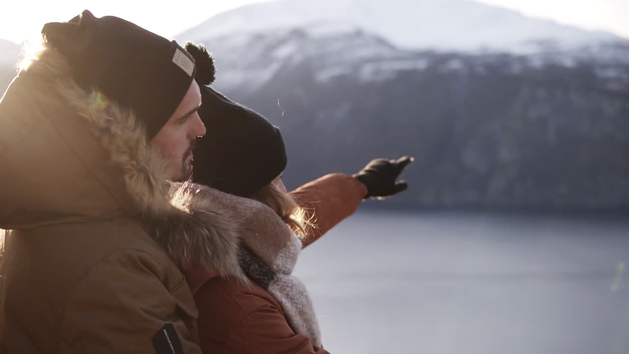 Young couple in winter coats embracing, people hugging looking at stunning view on lake and mountains peaks. Admire Beautiful Mountain Nature Landscape. Side view