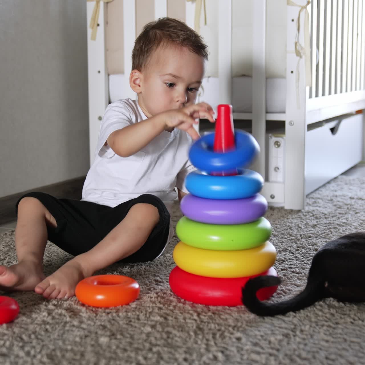 Baby boy building toy pyramid sitting on the floor. Black cat lies calm beside waving his tail