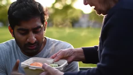 a young man sits in a park as another person hands him a steaming container of food. the warm evening light creates a relaxed atmosphere.