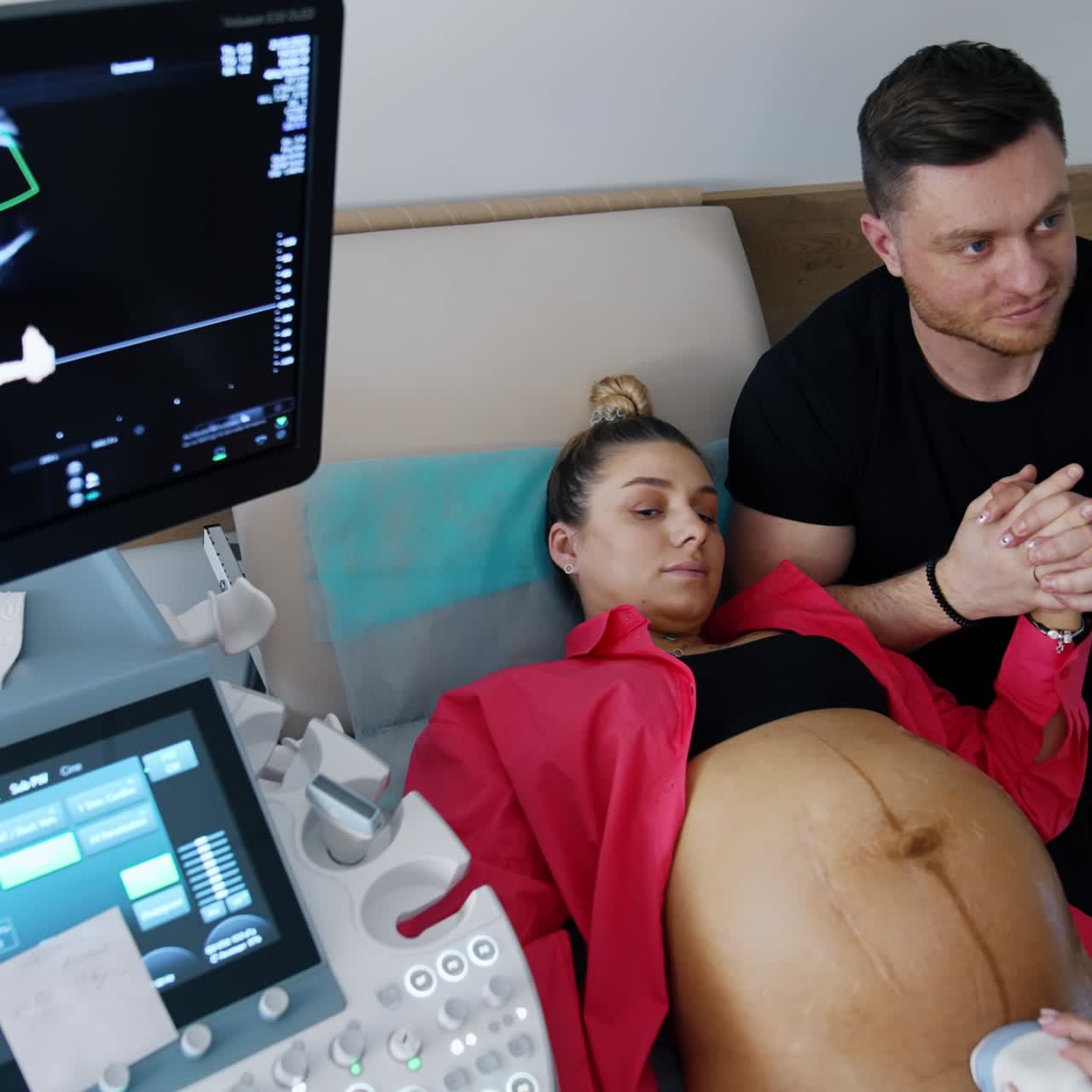 Woman expecting a baby lies on couch examined by the doctor on ultrasound machine. Man sits holding his wife's hand and looking on the screen. Top view