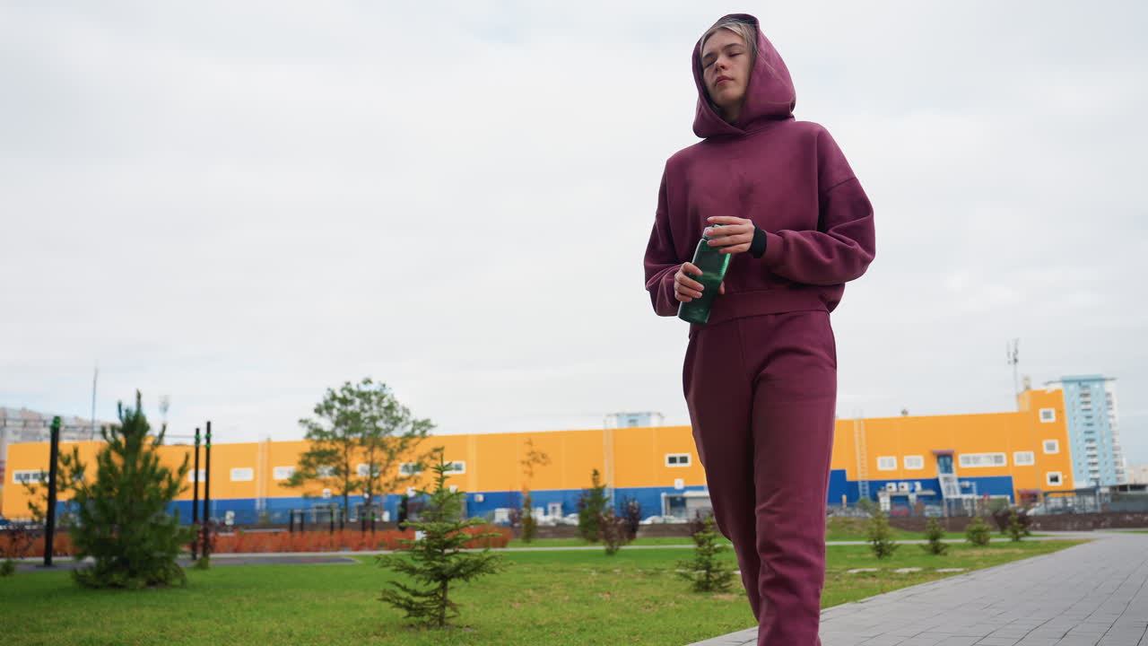 Young sportswoman in burgundy hoodie and matching joggers holds green water bottle while strolling along urban path lined with orange autumn foliage and tall apartment buildings under cloudy sky