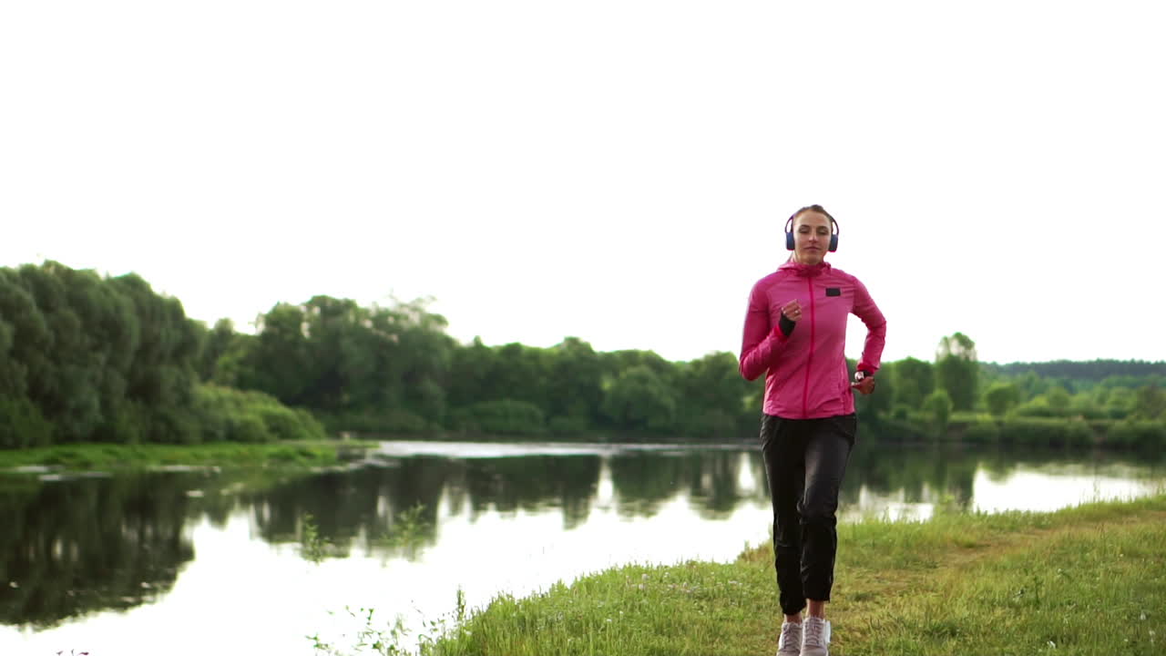 morena con cabello largo en auriculares corre a lo largo del río en el parque por la mañana al amanecer en el verano en una chaqueta rosa y pantalones negros