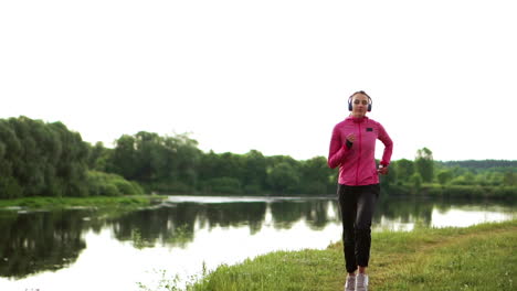 Brunette-with-long-hair-in-headphones-runs-along-the-river-in-the-Park-in-the-morning-at-sunrise-in-the-summer-in-a-pink-jacket-and-black-pants