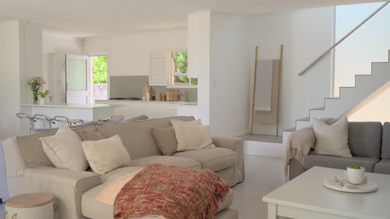 Kamera bergerak perlahan ke kanan, memperlihatkan sofa sectional krem dengan selimut merah, menunjukkan tata letak rencana terbuka yang dijual.