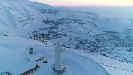 Rotación-De-Drones-Sobre-La-Estatua-De-San-Charbel-En-Un-Paisaje-Montañoso-Nevado-Al-Atardecer