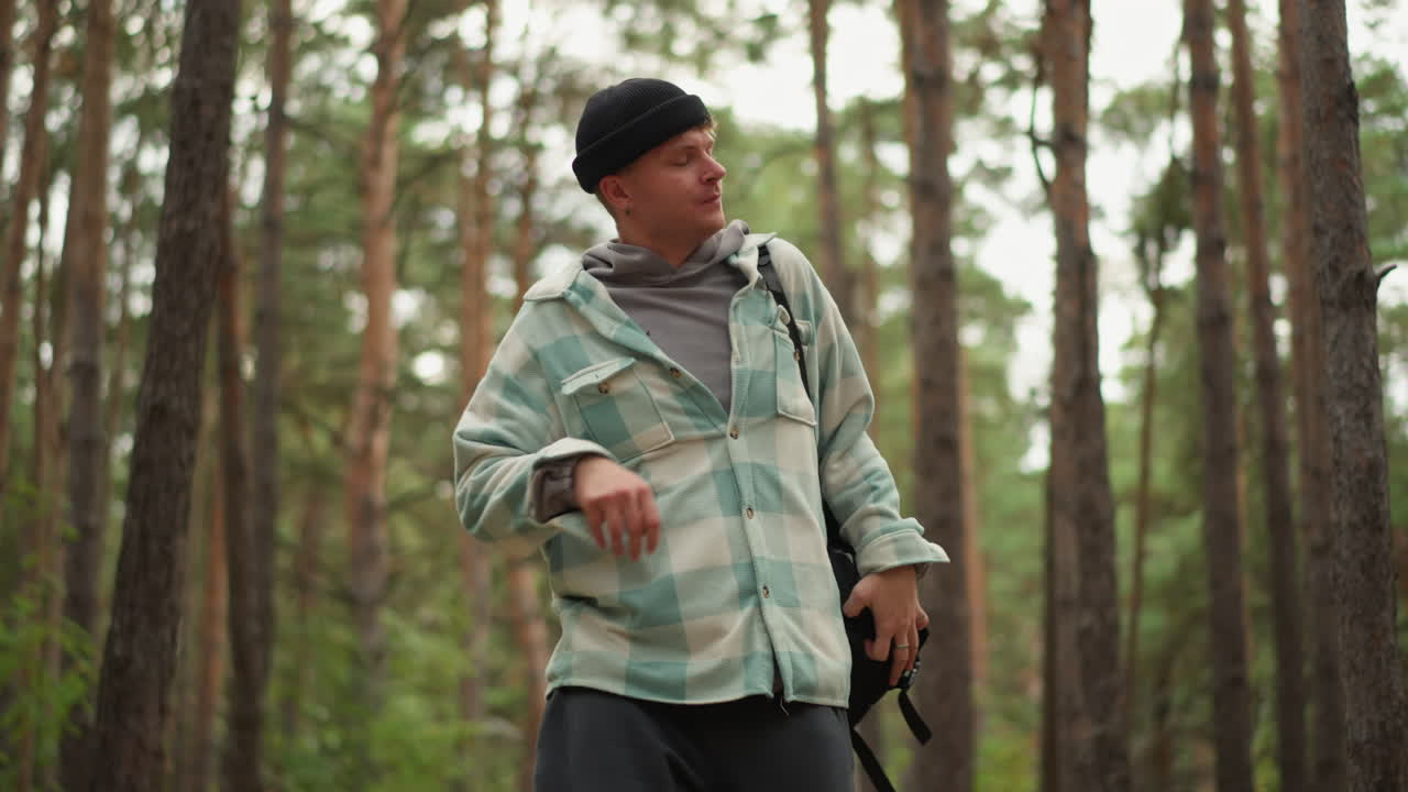 young man hiking through pine forest adjusting backpack and checking straps, wearing beanie and checkered jacket, steady stride among tall trunks, overcast light creating calm exploration mood